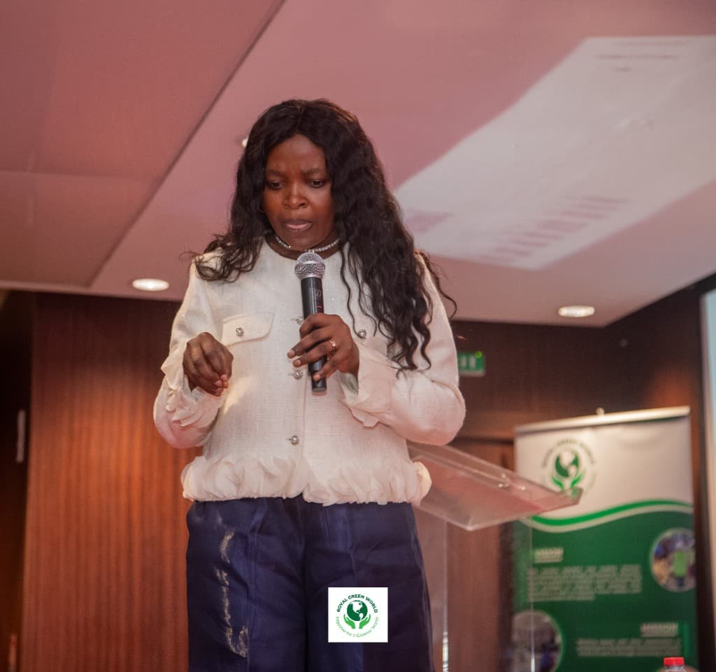 Speaker in a white jacket delivering remarks from a clear podium, with a Royal Green World banner behind her